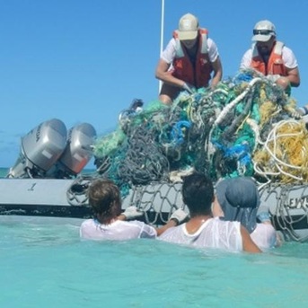 3 people floating in the water next to a boat helping the boat crew pull out a net full of garbage from the teal blue ocean